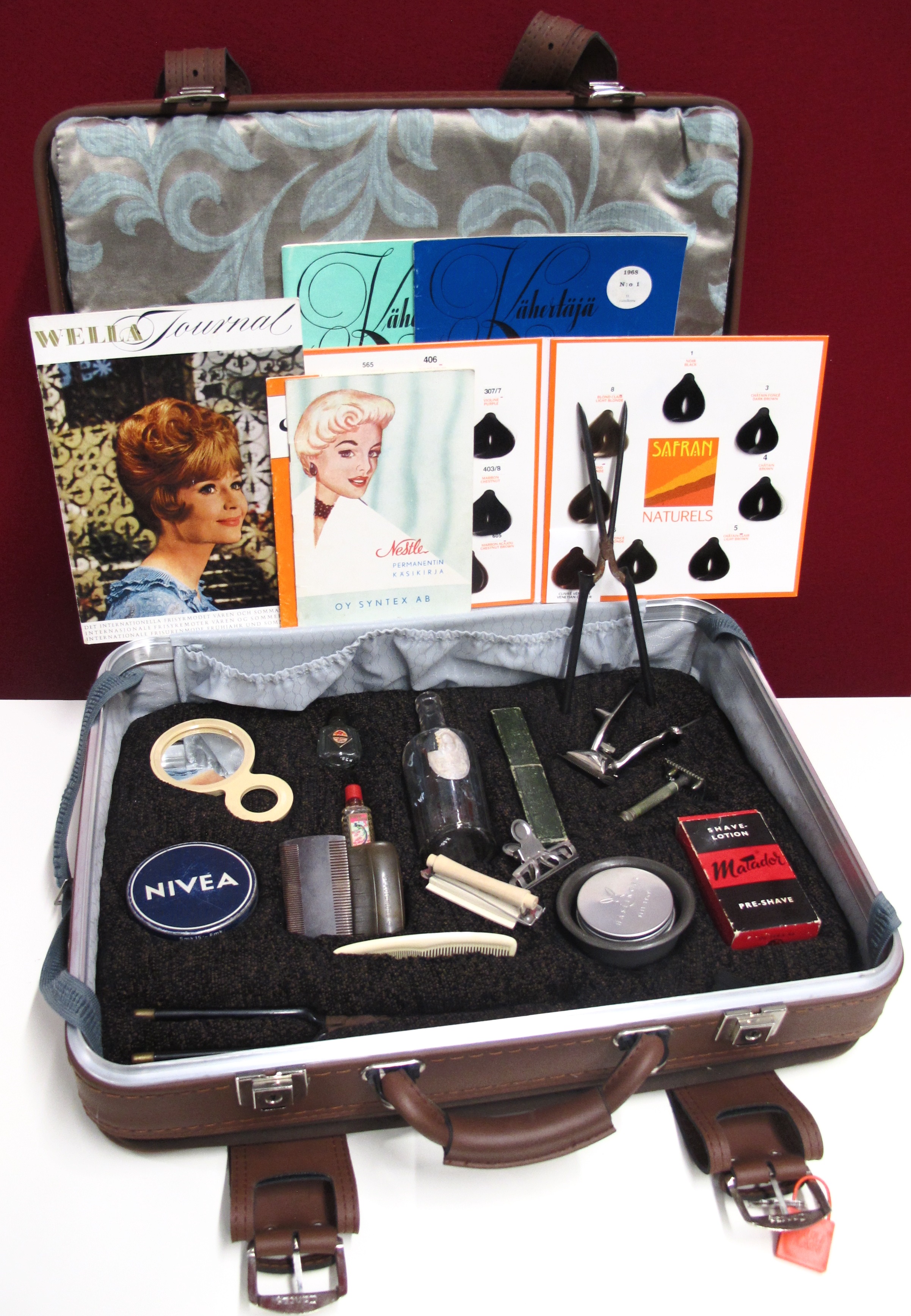 Vanha matkalaukku, jonka sisällä kauneudenhoitoon liittyviä lehtiä ja esineitä kuten erilaisia kampoja ja erikokoisia pulloja, purkkeja ja peili